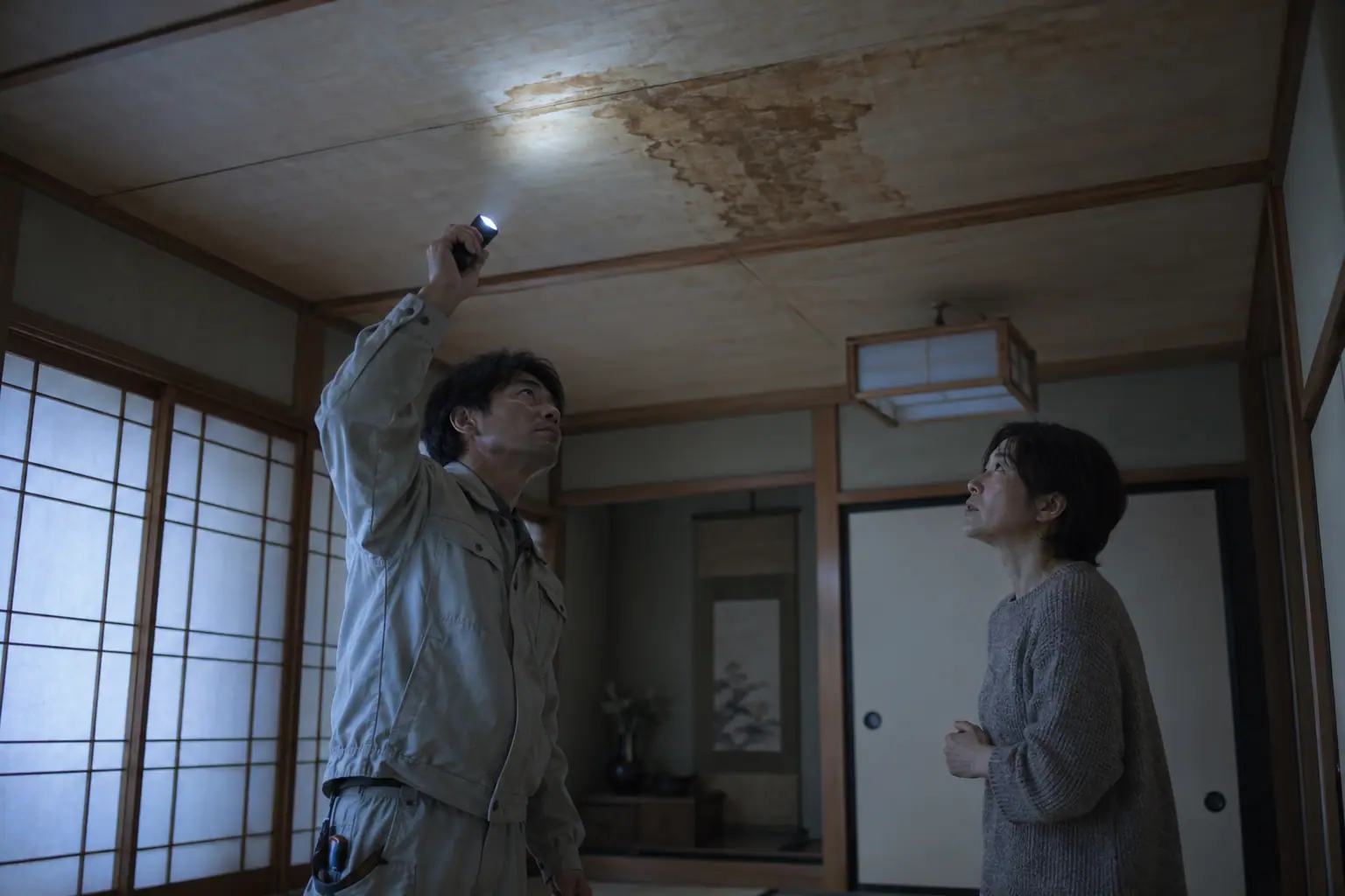 一戸建ての住宅の2階の和室で、職人が天井の雨染みを懐中電灯で照らして確かめている